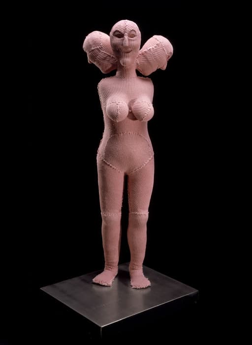 A sculptural figure of a standing female form covered entirely in pale pink knit fabric. The figure has exaggerated breasts, a slightly protruding belly, and rounded hips. Distinct stitching and seams are visible at the joints and torso. The head features hollow eye sockets and large rounded protrusions resembling stylized pigtails or ear-like shapes on either side. The figure stands on a square black pedestal against a completely black background, emphasizing its texture and form.
