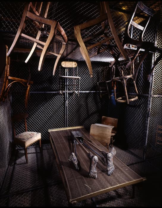 A dark, enclosed room with chain-link fencing on all sides, forming a cage-like environment. Various mismatched wooden chairs are suspended upside-down from the ceiling, creating a disorienting atmosphere. In the center of the space sits a metal mesh table with four sculptural, animal-like feet attached to its surface. Additional chairs of different styles are scattered around the room, some positioned normally on the floor. The lighting emphasizes the textures of the metal fencing and the worn wood of the furniture, contributing to an unsettling, surreal mood.