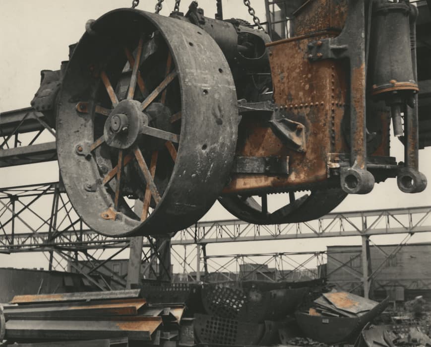 Close-up photograph of a large, rusted industrial wheel suspended by chains at a scrap yard. The heavy machinery part hangs mid-air, revealing bolts and worn surfaces. Below it, stacks of metal sheets and disassembled parts are scattered across the ground, with steel framework structures in the background.