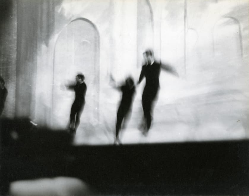 Blurry black-and-white photograph of four figures in motion on a stage. The performers, dressed in dark clothing, appear to be mid-jump or dance against a light-toned backdrop with arched shapes, creating a ghostly, abstract effect.