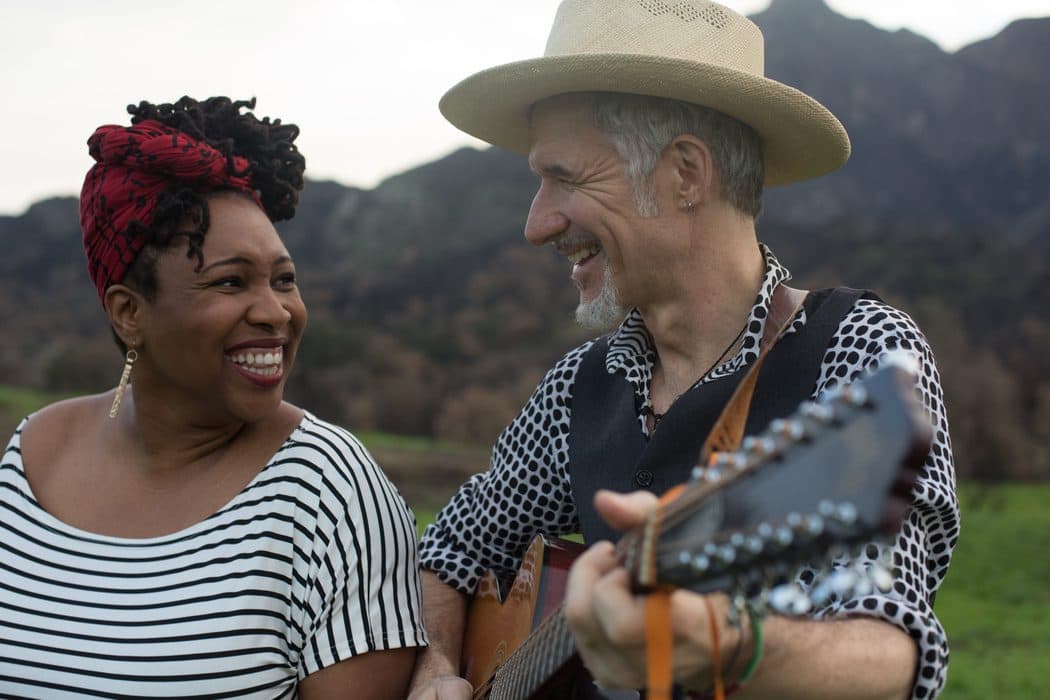 Smiling man and woman outdoors, with the man playing guitar and wearing a straw hat, and the woman wearing a striped shirt and red headwrap, standing against a blurred mountain backdrop.