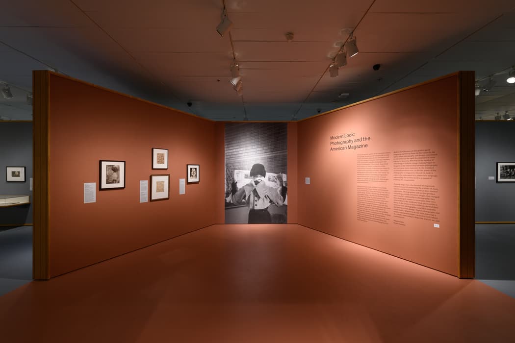 Exhibition gallery view with rust-colored walls from the show Modern Look: Photography and the American Magazine. The central wall features a large black-and-white photograph of a person reading a newspaper, standing in front of a tall modernist building. The left wall displays a selection of framed photographs and texts, while the right wall contains the exhibition introduction text. The surrounding galleries, visible in the background, have gray-blue walls with additional framed artworks. The floor and ceiling lighting accentuate the clean, modern presentation.