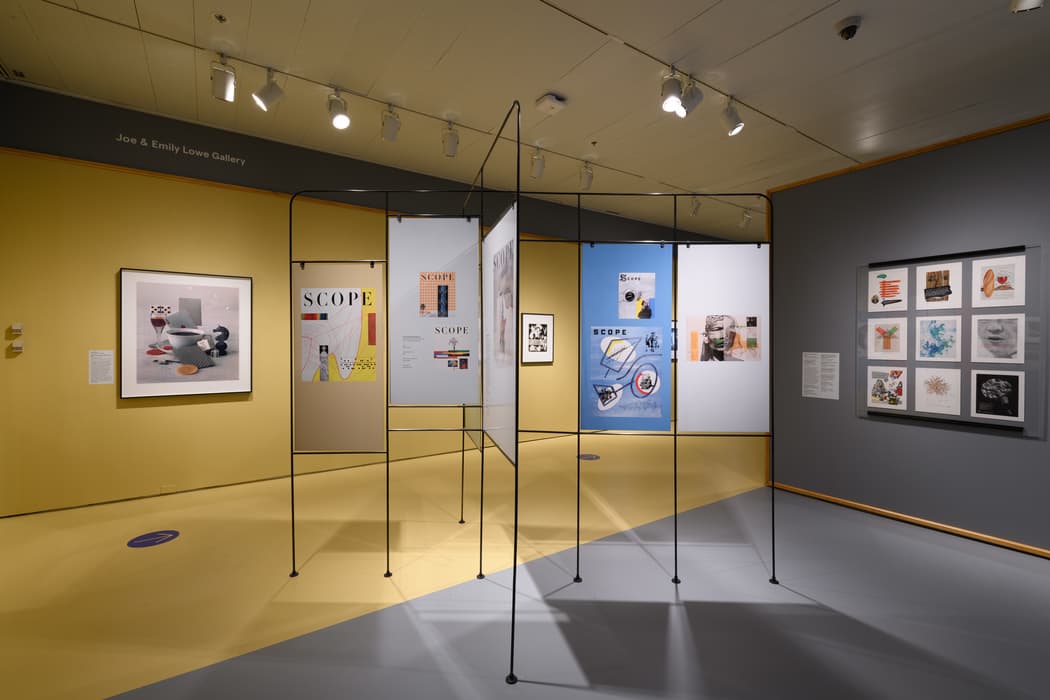 Gallery view from the exhibition Modern Look: Photography and the American Magazine. The room features bright yellow walls and floor with gray accents. In the center, a metal framework structure holds mounted posters and magazine spreads from Scope, showcasing modern graphic design and photography. Additional framed works are displayed on the surrounding walls, including abstract and photographic compositions. Overhead track lighting illuminates the works, while the clean, geometric layout highlights the modernist design aesthetic.