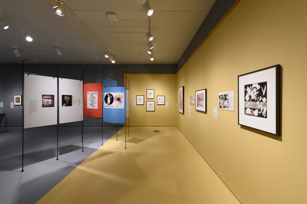 Gallery view from Modern Look: Photography and the American Magazine. The space features yellow walls and floors with gray accent walls. On the left, a freestanding metal framework displays mounted prints and magazine pages, including covers from Scope and a 