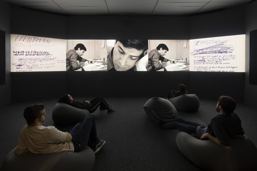 Visitors seated on beanbags watch a panoramic projection of Leonard Cohen writing in a notebook, surrounded by handwritten notes.