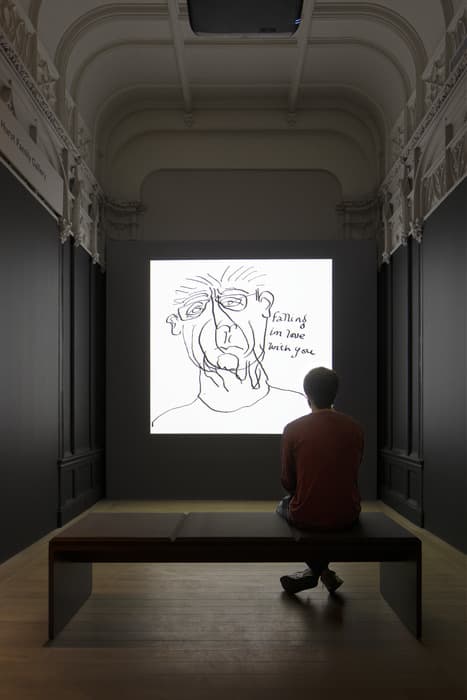 Person seated on a bench viewing a projection of a line-drawn portrait with the handwritten text “Falling in love with you” in a dimly lit gallery.