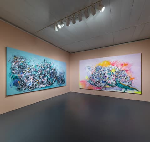 Gallery space with soft peach-colored walls displaying two large abstract paintings. The left painting features swirling blue, gray, and pink forms against a light blue background, while the right painting shows a similar composition with brighter pink, yellow, and green tones against a pale pink background. Overhead track lighting illuminates both artworks.
