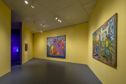 Gallery space with bright yellow walls displaying colorful contemporary paintings. The center painting features abstracted human figures in red, yellow, and purple tones on a dark background. The painting on the right shows layered imagery in vibrant reds, blues, and yellows. Overhead lighting highlights each artwork, with a glimpse into an adjacent purple-lit room.