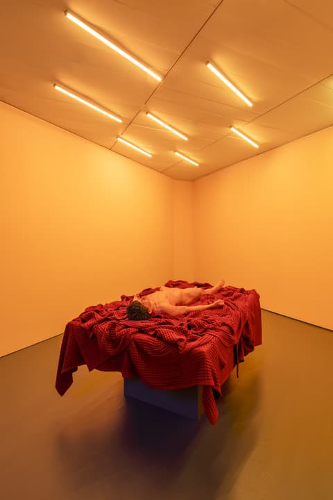Installation view of a gallery with warm yellow lighting and a ceiling of fluorescent tube lights. In the center is a sculpture of a nude figure lying face up on a platform covered in red fabric.