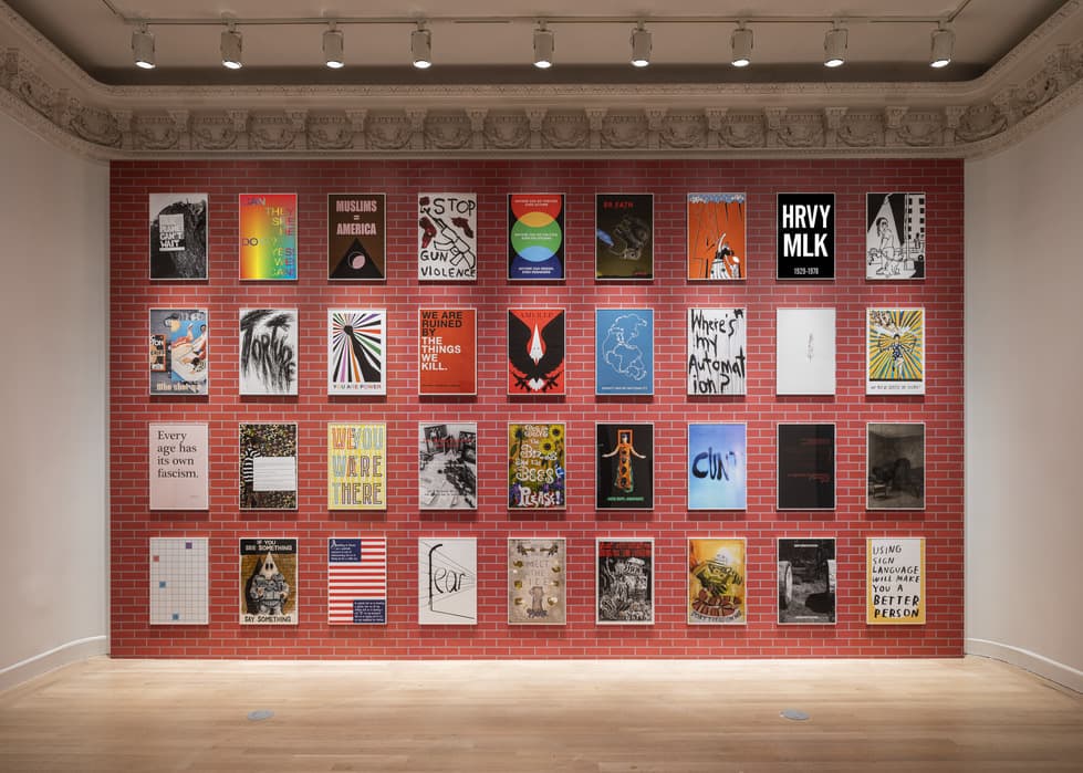 Gallery wall covered in red brick wallpaper, displaying five rows of colorful, graphic posters with political and social messages, under ceiling track lighting.