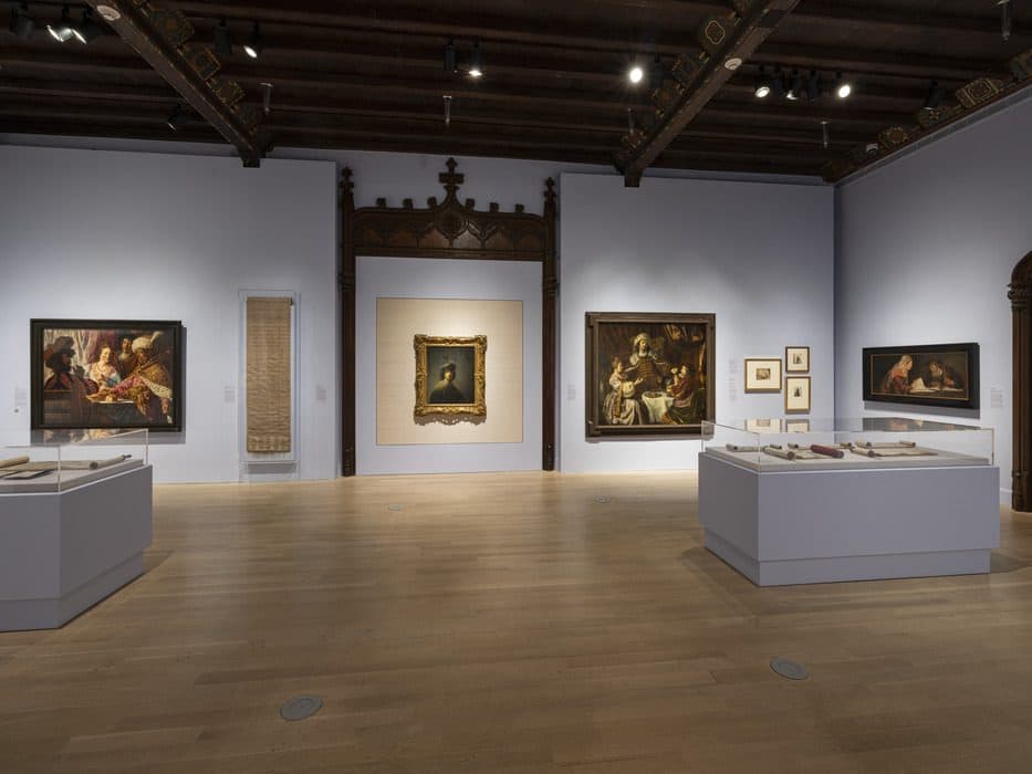 Museum gallery displaying framed paintings, scrolls, and historical texts in glass cases, with light blue walls, wood flooring, and ornate woodwork framing the central artwork.