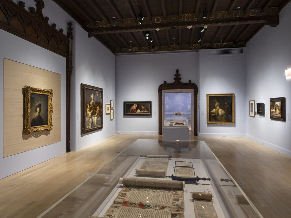 Gallery view of the Book of Esther exhibition featuring framed paintings on the walls, historical manuscripts displayed in a glass case, and ornate wood architectural details.