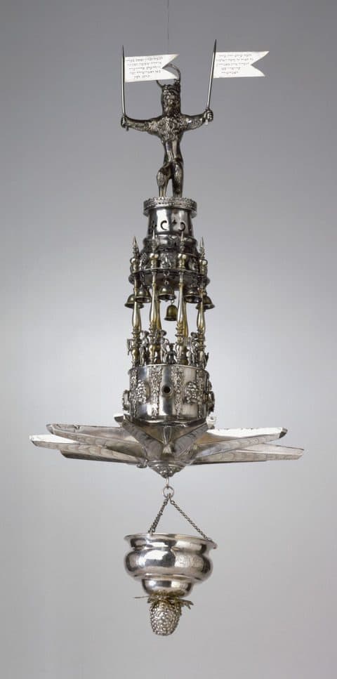 Ornate silver hanging spice container with a human figure holding two flags at the top, multiple bells, decorative figures and grape motifs, and a suspended vessel with a pinecone-shaped finial at the bottom.