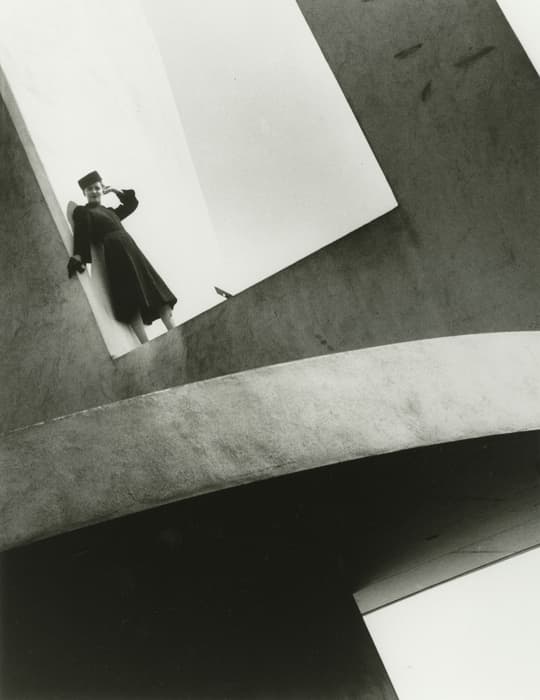 Person wearing a long dark coat and hat stands in a high angular opening of a modernist concrete structure, viewed from below. The geometric architecture features curved and slanted planes with dramatic contrast between light and shadow.