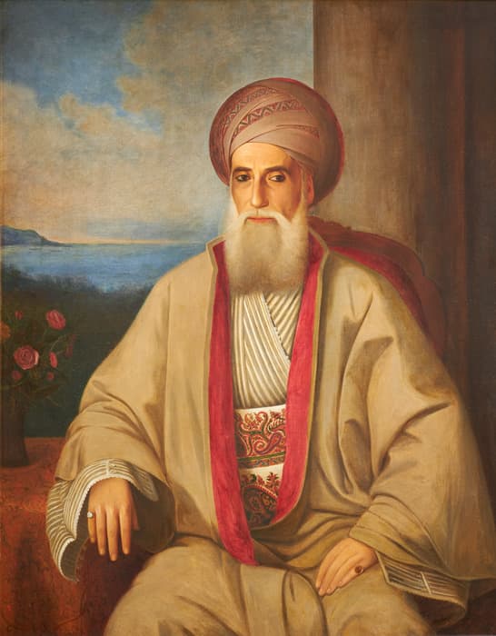 Portrait painting of a seated man wearing traditional Middle Eastern attire, including a tan robe with a red trim, a patterned sash, and a tall, wrapped turban. He has a long white beard and looks directly at the viewer with a calm expression. His right hand rests on an ornate table with a vase of pink roses, while the left hand rests on his lap. In the background is a scenic landscape with a body of water and distant hills under a cloudy sky.