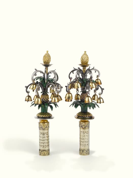 A pair of ornate Torah finials (rimmonim), crafted from metal and adorned with small golden bells. Each finial features a central column with Hebrew inscriptions on a cylindrical base, topped by elaborate branches resembling palm trees with gilded pomegranate shapes and leaves. The tops are crowned with gold pinecone-like ornaments. The objects have green, gold, and silver tones and are set against a plain, light background.
