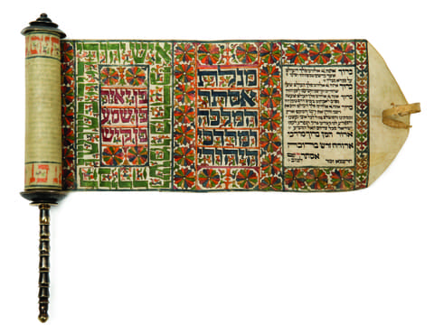 An illuminated scroll partially unrolled, with a turned wooden handle on the left. The scroll features three distinct vertical panels: the left and center panels display colorful geometric floral designs in red, green, and orange, framing large Hebrew text in white and black. The right panel contains blocks of smaller Hebrew text in black ink on a parchment background. Decorative floral borders run along the top and bottom of the scroll, enhancing its intricate design.