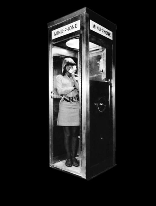 Black-and-white image of a woman standing barefoot inside a narrow phone booth labeled 