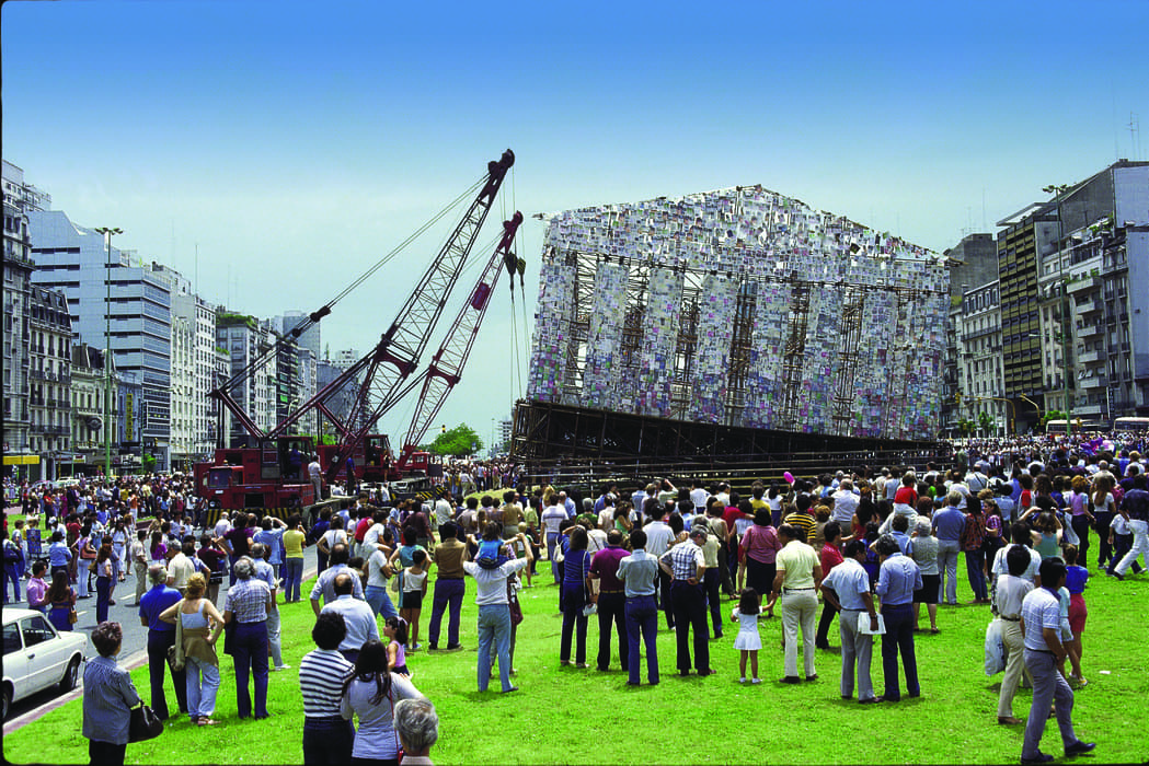 Large crowd gathers in a city plaza to watch cranes installing a massive temporary structure resembling a classical building, constructed from panels covered with books or printed pages, surrounded by tall urban buildings under a clear blue sky.