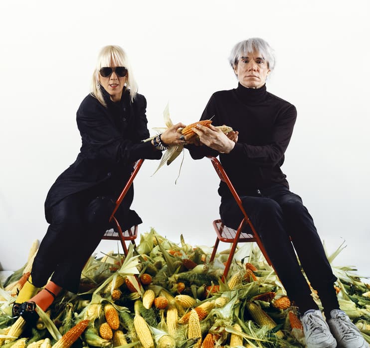 Two people sit on red chairs amid a large pile of corn. The woman on the left wears black clothing and sunglasses; the man on the right wears a black turtleneck and light sneakers. Both hold an ear of corn together, extending it toward the camera.