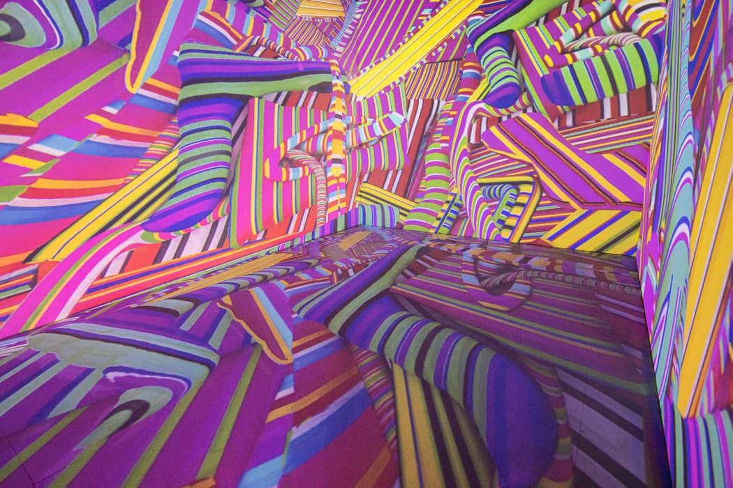 Immersive installation space filled with vibrant, overlapping, multicolored stripes in shades of purple, pink, yellow, green, blue, and red, creating an optical illusion of twisting, undulating forms across the walls, floor, and ceiling.