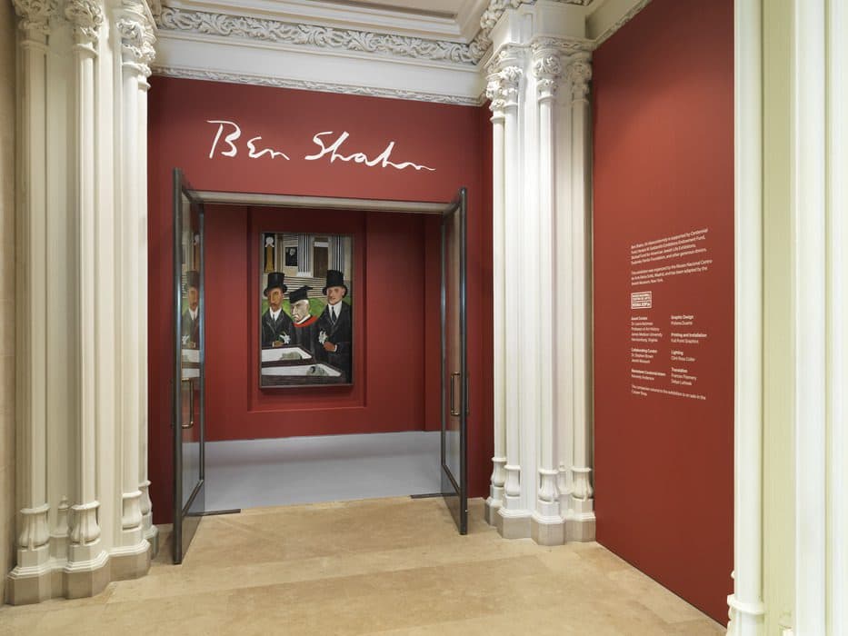 Entrance to the Ben Shahn exhibition at the Jewish Museum, with red walls, white architectural columns, and a painting of three men in suits and top hats visible through open doors.