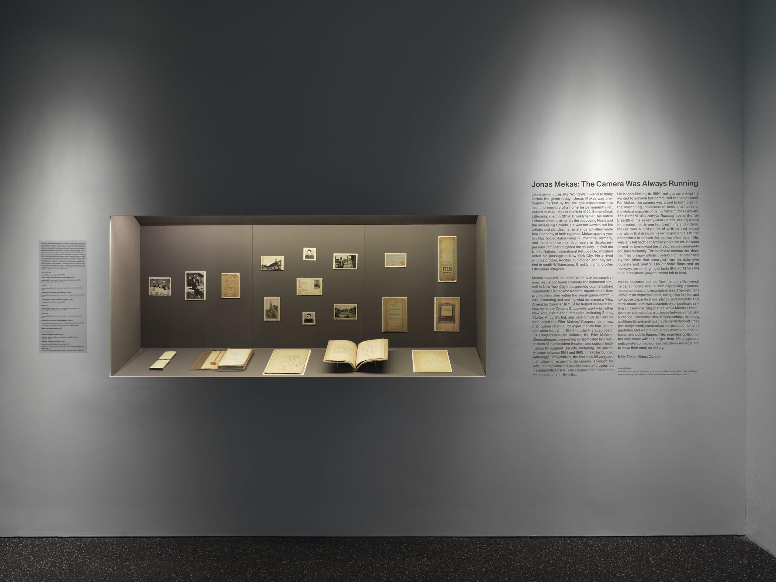 Exhibit wall with a display case containing photographs, documents, and open books related to Jonas Mekas. On the right wall, exhibition text titled “Jonas Mekas: The Camera Was Always Running” provides biographical context.