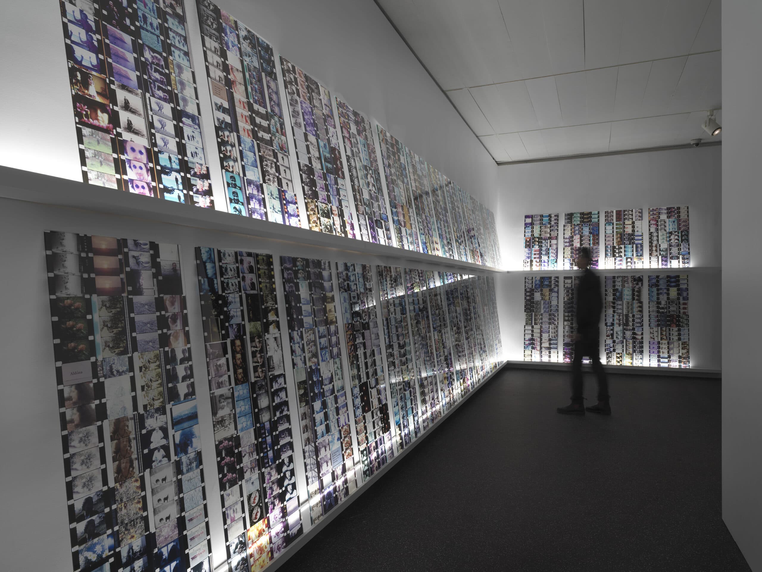 Gallery room with walls covered in large vertical strips of film frames arranged in dense grids. A person stands in the room viewing the installation, which is dramatically lit from below and behind the film panels.