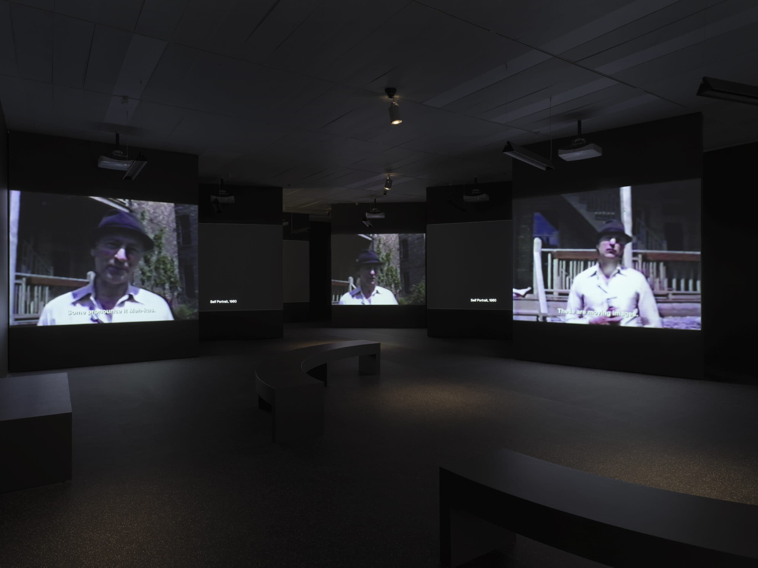 Dark gallery with multiple projection screens showing video footage of a man in a white shirt and dark hat standing outdoors. Subtitles and the text “Self Portrait, 1980” appear on some screens. Curved black benches are in the center of the room.