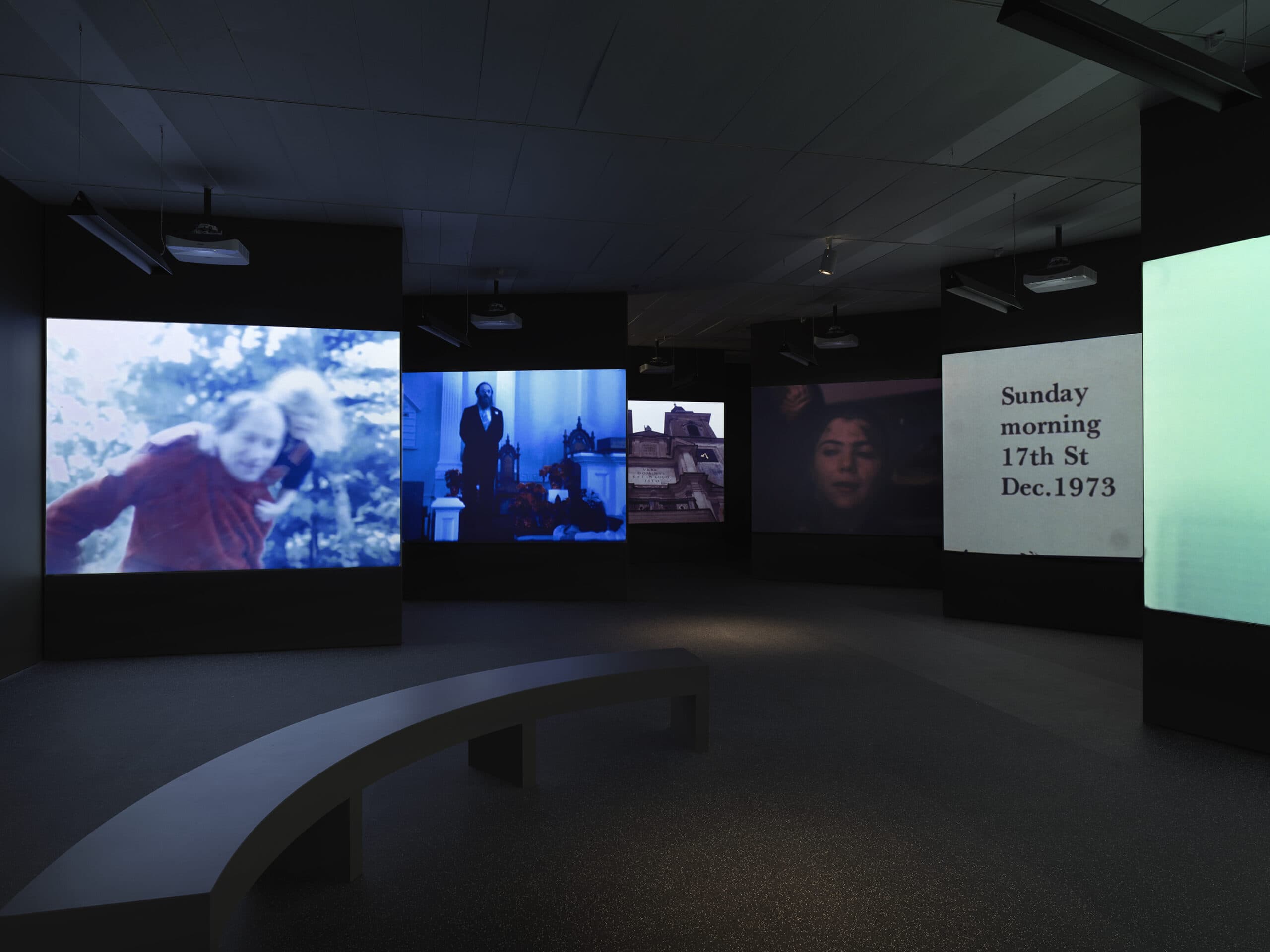 Dark gallery space with multiple projection screens showing different video scenes, including a man carrying a child, a man standing at a church altar, a woman's face, and a screen with the text “Sunday morning 17th St Dec. 1973.” Curved black benches are positioned in the center of the room.
