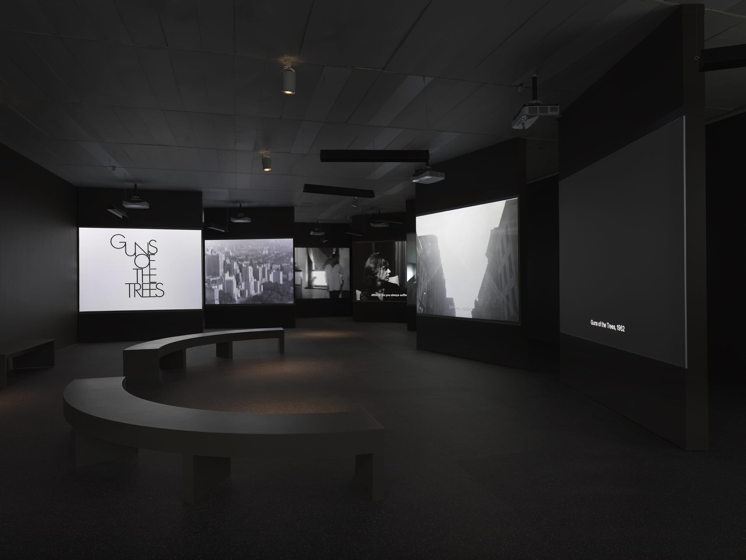 Dark gallery space featuring multiple large projection screens showing scenes from Guns of the Trees (1962). The left screen displays the film title in black text on a white background. Other screens show black-and-white urban and portrait imagery. Curved black benches are placed in the foreground.