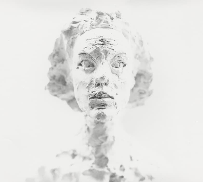 Close-up photograph of a sculpted bust of a woman, rendered in a highly textured, rough style. The white sculpture shows a solemn face with wide, expressive eyes, detailed lips, and softly indicated hair, set against a stark white background that blends into the figure's shoulders and neck.