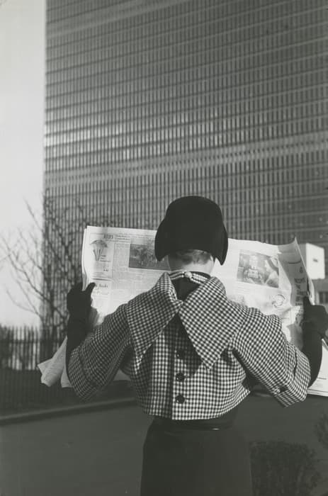 Person wearing a checkered coat and dark hat stands outdoors reading a newspaper, facing a tall modernist glass building. The figure is seen from behind, with gloved hands holding the open newspaper.