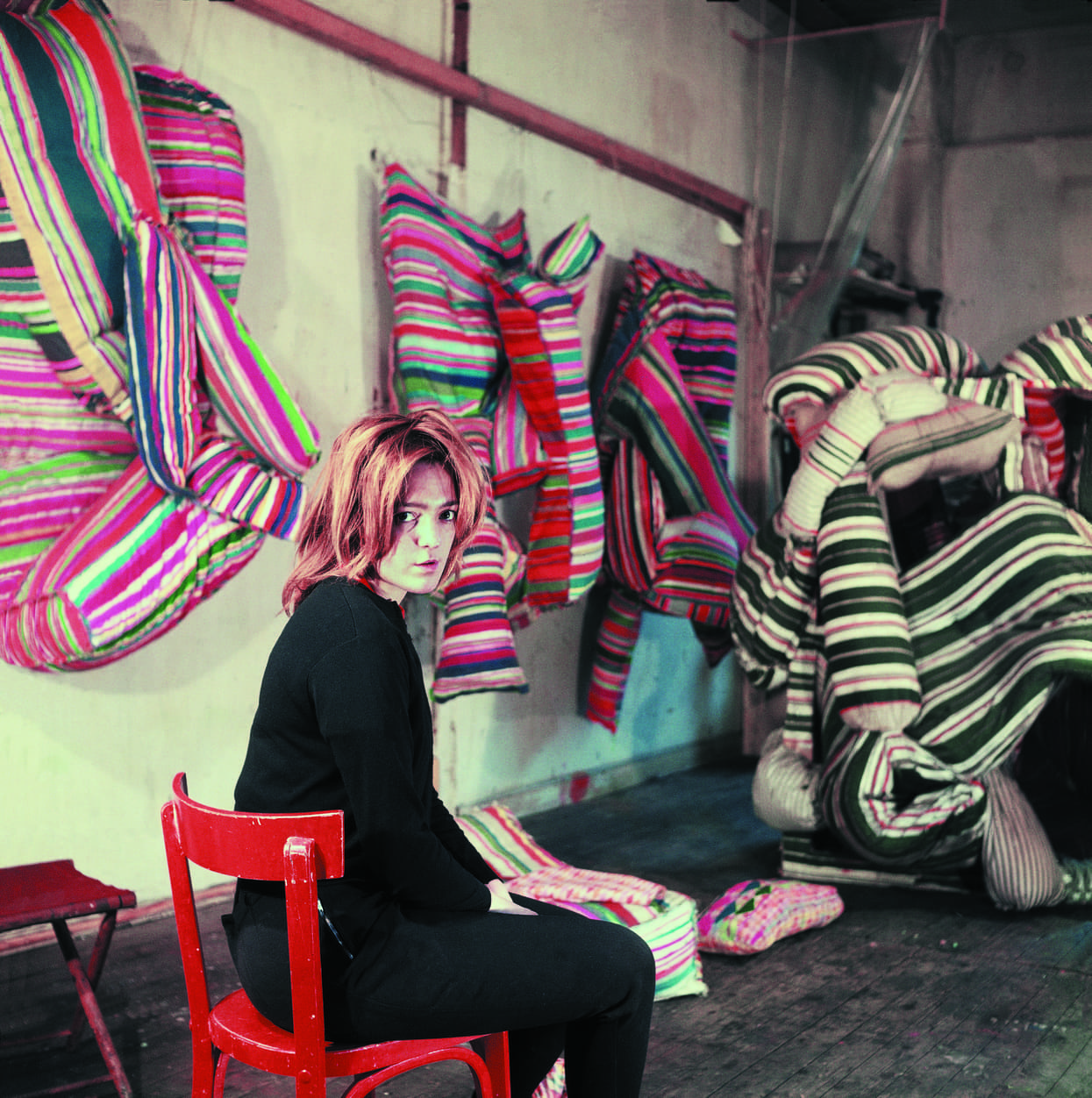 Artist seated on a red chair in front of colorful, striped fabric sculptures hanging and piled in a studio space with wooden floors and a beige wall.