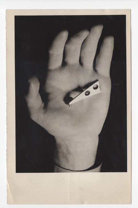 Black-and-white photograph of an open palm facing the viewer, holding a small, abstract metal object with two circular holes. The hand is well-lit against a dark background, with high contrast emphasizing the object and the hand’s texture. The image is framed by a white border with slight wear on the edges.