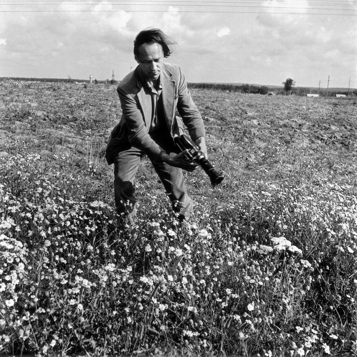 Black and white photo of a man in a field of wildflowers, bending forward while holding a film camera.