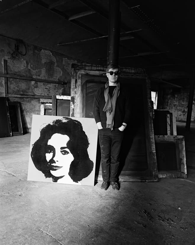 Evelyn Hofer. Andy Warhol standing in the factory with a Liz painting and several screens in the background, January 29, 1964. Image provided by the estate of Evelyn Hofer.