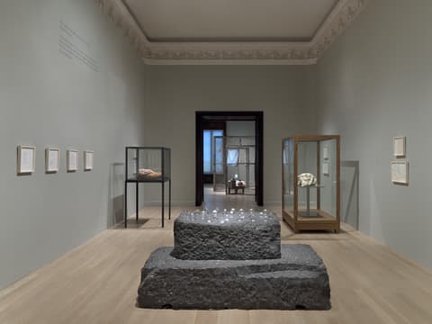 A museum gallery with pale gray walls and light wood floors. In the center, a large two-tiered rectangular sculpture made of rough gray stone is positioned on the floor, with small glass objects placed on its top surface. To the left, a black-framed glass case displays a sculpture of a pale pink reclining figure. To the right, a wooden-framed glass case contains a white organic-shaped sculpture. Framed works on paper hang on the walls surrounding the sculptures. In the background, an open doorway reveals another gallery space with large windows and additional artwork. The gallery features ornate crown molding and soft, even lighting.