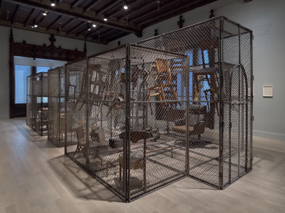 A museum gallery displaying a large sculptural installation consisting of several connected chain-link enclosures. Inside the enclosures, numerous wooden chairs and tables are suspended upside down or at odd angles, creating a chaotic, disorienting scene. The room features high ceilings with dark wooden beams, ornate carved woodwork around the windows, and pale wooden floors. Soft lighting and natural light from the tall windows illuminate the installation.