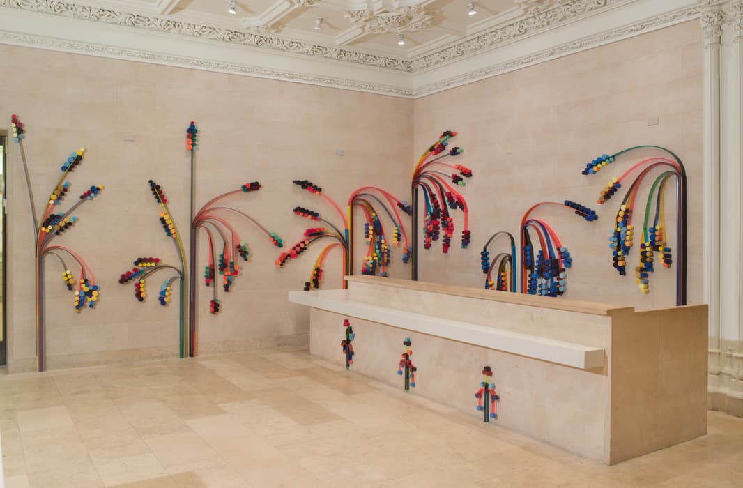 Colorful sculptural installation of flexible tubing and foam cylinders arranged in playful, palm-like bursts on the walls and desk of a beige marble room with ornate ceiling moldings.