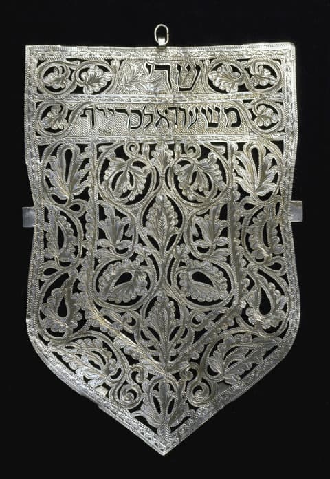Intricately crafted silver Torah shield with an openwork floral and foliate design, Hebrew inscription across the top, and symmetrical decorative patterns throughout, set against a black background.