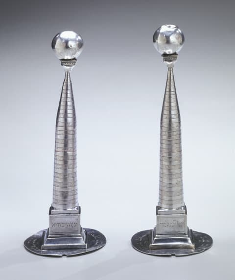 Pair of tall, slender silver Torah finials shaped like tapering columns, each topped with a polished silver sphere. The bases are square with Hebrew inscriptions, resting on round flat plates.