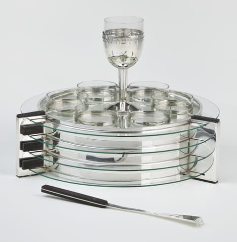 Modernist silver and glass Passover seder plate with multiple stacked circular glass tiers. On top sit eight small glass dishes arranged in a circle with one central goblet. A silver serving utensil with a black handle lies in front of the plate.