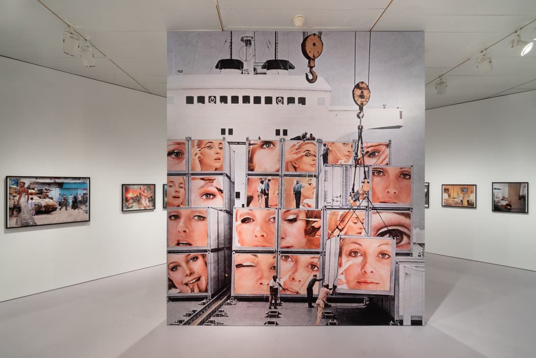 Gallery installation featuring a large central artwork showing a photomontage of shipping containers stacked on a cargo ship, each container displaying close-up images of women's faces and eyes. The surrounding gallery walls display additional photographic artworks in black frames.