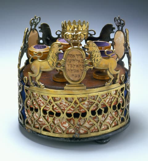 Ornate ceremonial object resembling a crown, featuring an openwork metal lattice design with lions, a crown at the top, Hebrew inscription, and small cups arranged inside on a wooden base.