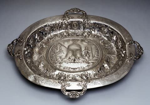 Ornate oval silver basin with raised handles and elaborate floral designs around the border; the center depicts a tented structure with two human figures, surrounded by detailed engraved foliage and patterns.