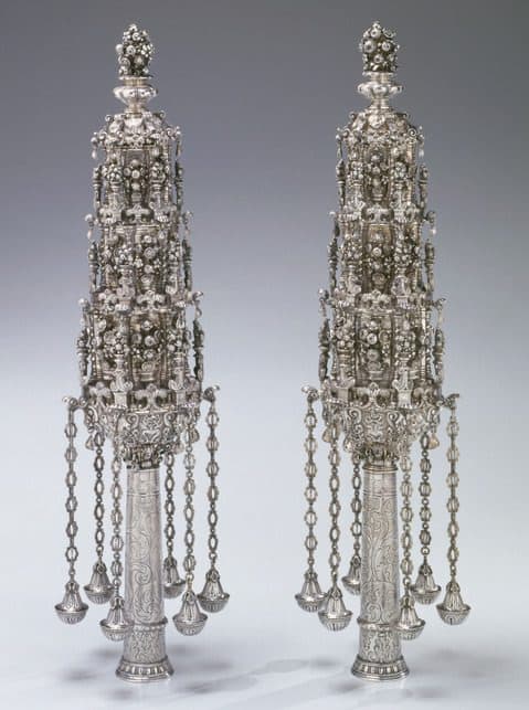 Pair of ornate silver Torah finials with multiple tiers of intricate floral designs, bells hanging from chains, and detailed engraving on the stems, displayed against a neutral background.