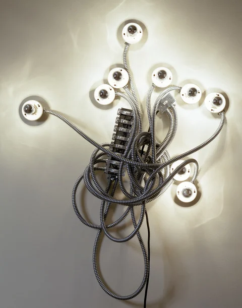Wall-mounted light sculpture with exposed silver electrical conduits extending in multiple directions, ending in nine lit bulbs, resembling an abstract menorah or branching form, set against a plain wall.