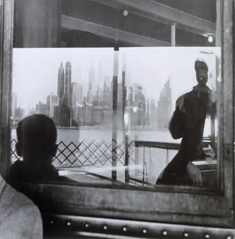 View of Manhattan skyline reflected in a ferry window, with silhouetted passengers and metal railing in foreground.