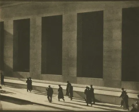 Sepia-toned photograph of pedestrians walking along a city sidewalk in front of a large stone building with tall rectangular dark windows, casting elongated shadows across the scene.